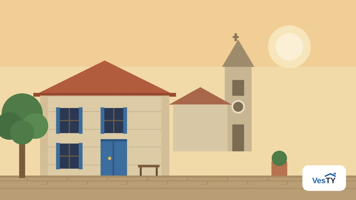 Une maison de bourg à façade de pierre et volets bleus, un arbre et un banc devant elle, le clocher d'une église en arrière-plan dans la lumière dorée de fin de journée